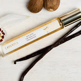 Lavanila Pure Vanilla rollerball perfume with gold cap arranged on pale wood beside vanilla pods, an orchid and tonka beans.