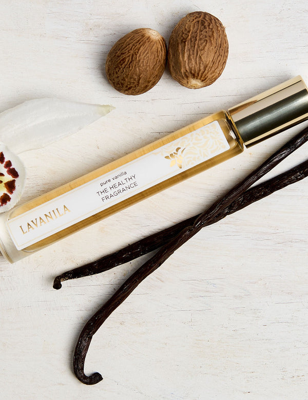 Lavanila Pure Vanilla rollerball perfume with gold cap arranged on pale wood beside vanilla pods, an orchid and tonka beans.