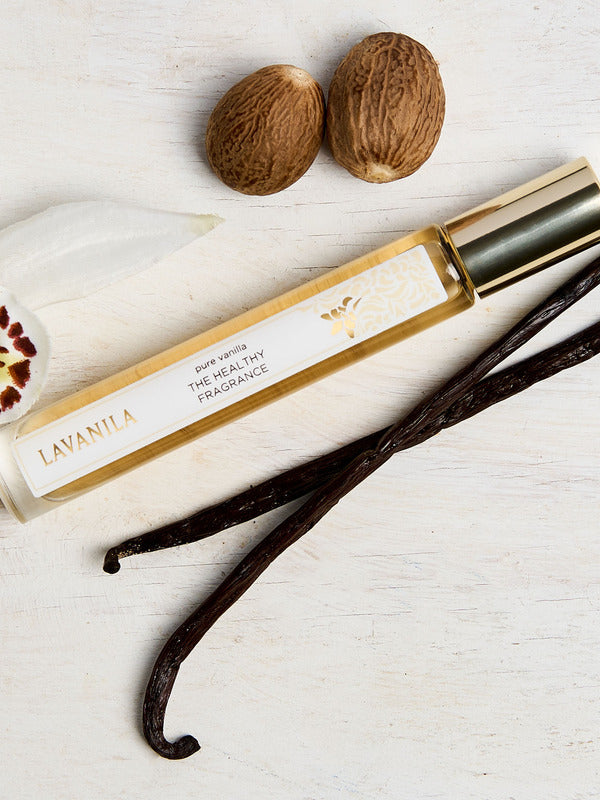 Lavanila Pure Vanilla rollerball perfume with gold cap arranged on pale wood beside vanilla pods, an orchid and tonka beans.