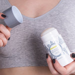 Person in a grey sports top holding an open Lavanila Sport Luxe High Performance Deodorant stick in one hand and its cap in the other.