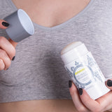 Person in a grey sports top holding an open Lavanila Sport Luxe High Performance Deodorant stick in one hand and its cap in the other.