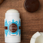 Open Lavanila Vanilla Coconut deodorant with a rounded white stick and turquoise botanical label, lying on dark wood beside coconut pieces and its brown cap.