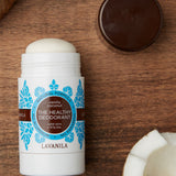 Open Lavanila Vanilla Coconut deodorant with a rounded white stick and turquoise botanical label, lying on dark wood beside coconut pieces and its brown cap.