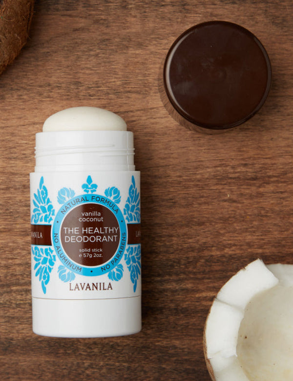 Open Lavanila Vanilla Coconut deodorant with a rounded white stick and turquoise botanical label, lying on dark wood beside coconut pieces and its brown cap.