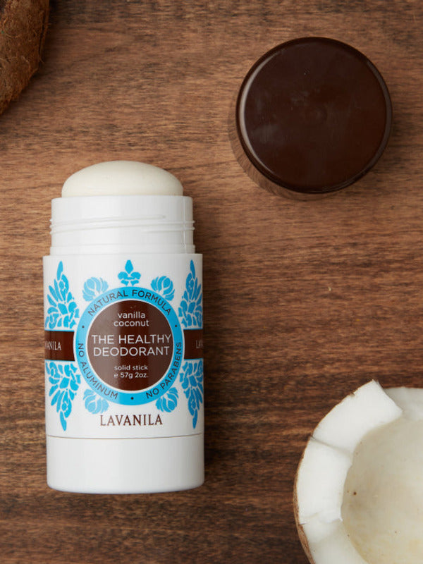Open Lavanila Vanilla Coconut deodorant with a rounded white stick and turquoise botanical label, lying on dark wood beside coconut pieces and its brown cap.