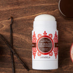Open Lavanila Vanilla Passionfruit deodorant with a rounded white stick and red-orange botanical label, lying on dark wood beside a vanilla pod and its brown cap.