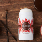 Open Lavanila Vanilla Passionfruit deodorant with a rounded white stick and red-orange botanical label, lying on dark wood beside a vanilla pod and its brown cap.