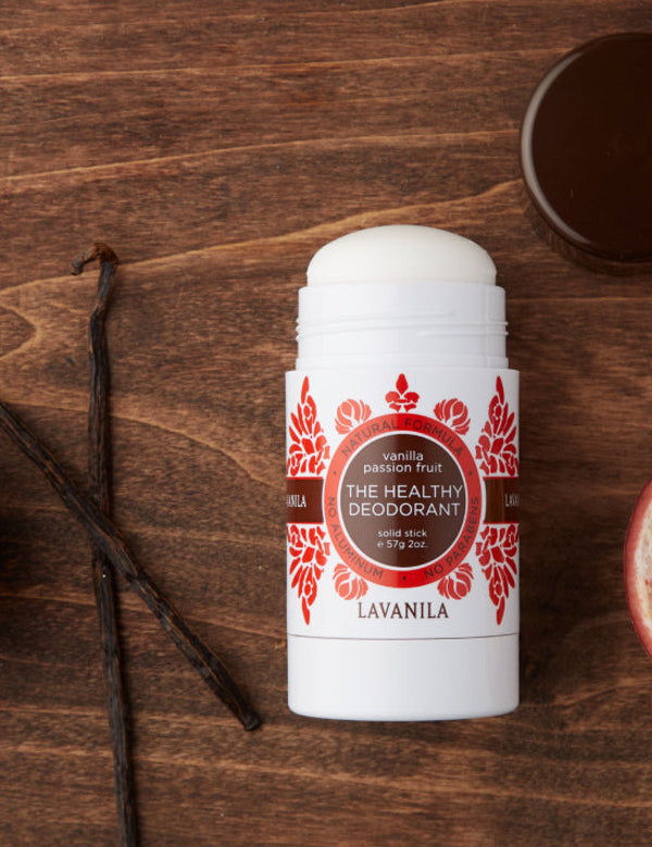 Open Lavanila Vanilla Passionfruit deodorant with a rounded white stick and red-orange botanical label, lying on dark wood beside a vanilla pod and its brown cap.