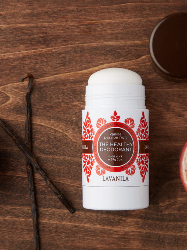 Open Lavanila Vanilla Passionfruit deodorant with a rounded white stick and red-orange botanical label, lying on dark wood beside a vanilla pod and its brown cap.