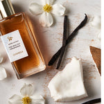 Rectangular Lavanila Vanilla Coconut perfume bottle with a gold cap lying diagonally on pale wood beside coconut pieces, dark vanilla pods and small white flowers with yellow centres.