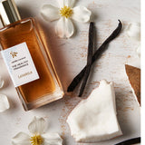 Rectangular Lavanila Vanilla Coconut perfume bottle with a gold cap lying diagonally on pale wood beside coconut pieces, dark vanilla pods and small white flowers with yellow centres.