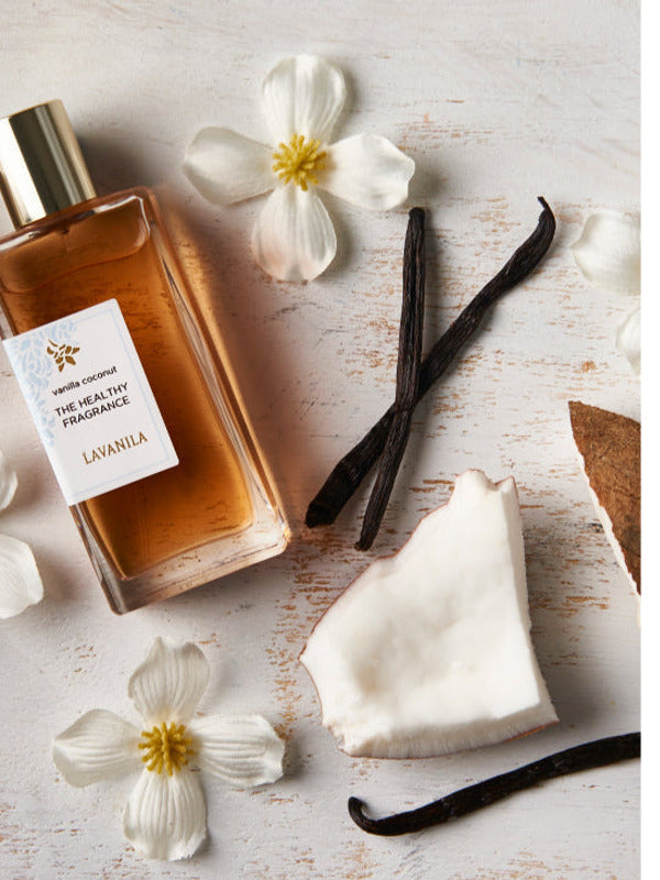 Rectangular Lavanila Vanilla Coconut perfume bottle with a gold cap lying diagonally on pale wood beside coconut pieces, dark vanilla pods and small white flowers with yellow centres.
