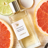 Rectangular Lavanila Vanilla Grapefruit perfume bottle with a gold cap lying diagonally on pale wood among pink grapefruit halves, lime slices, white petals and dark vanilla pods.