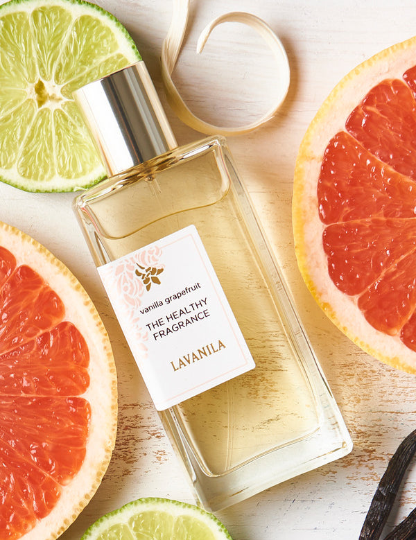 Rectangular Lavanila Vanilla Grapefruit perfume bottle with a gold cap lying diagonally on pale wood among pink grapefruit halves, lime slices, white petals and dark vanilla pods.