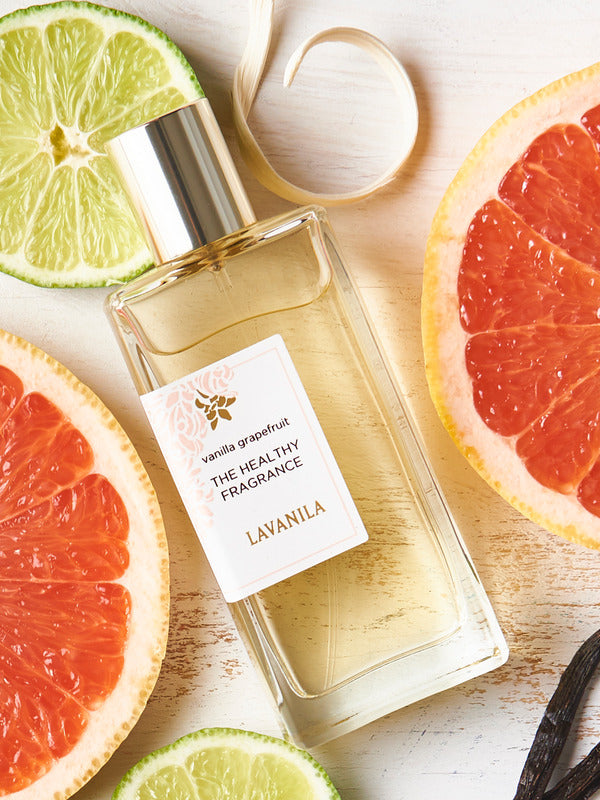 Rectangular Lavanila Vanilla Grapefruit perfume bottle with a gold cap lying diagonally on pale wood among pink grapefruit halves, lime slices, white petals and dark vanilla pods.