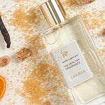Lavanila Vanilla Sugarcane perfume bottle with a gold cap lying diagonally among scattered golden sugar, brown sugar cubes, dark vanilla pods and an orange slice on pale wood.