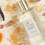 Lavanila Vanilla Sugarcane perfume bottle with a gold cap lying diagonally among scattered golden sugar, brown sugar cubes, dark vanilla pods and an orange slice on pale wood.