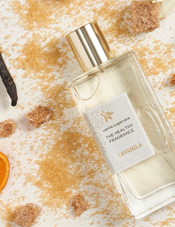 Lavanila Vanilla Sugarcane perfume bottle with a gold cap lying diagonally among scattered golden sugar, brown sugar cubes, dark vanilla pods and an orange slice on pale wood.