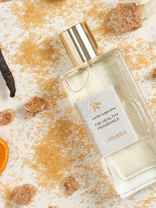 Lavanila Vanilla Sugarcane perfume bottle with a gold cap lying diagonally among scattered golden sugar, brown sugar cubes, dark vanilla pods and an orange slice on pale wood.