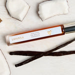 Slim Lavanila Vanilla Coconut rollerball perfume bottle with a gold cap lying on pale wood among pieces of white coconut flesh and dark vanilla pods.