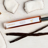 Slim Lavanila Vanilla Coconut rollerball perfume bottle with a gold cap lying on pale wood among pieces of white coconut flesh and dark vanilla pods.