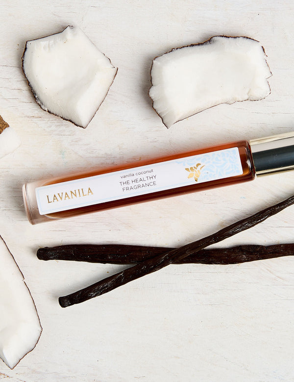 Slim Lavanila Vanilla Coconut rollerball perfume bottle with a gold cap lying on pale wood among pieces of white coconut flesh and dark vanilla pods.