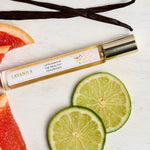 Slim Lavanila Vanilla Grapefruit rollerball perfume bottle with a gold cap lying diagonally on pale wood beside lime slices, pink grapefruit wedges and dark vanilla pods.