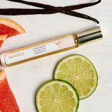 Slim Lavanila Vanilla Grapefruit rollerball perfume bottle with a gold cap lying diagonally on pale wood beside lime slices, pink grapefruit wedges and dark vanilla pods.