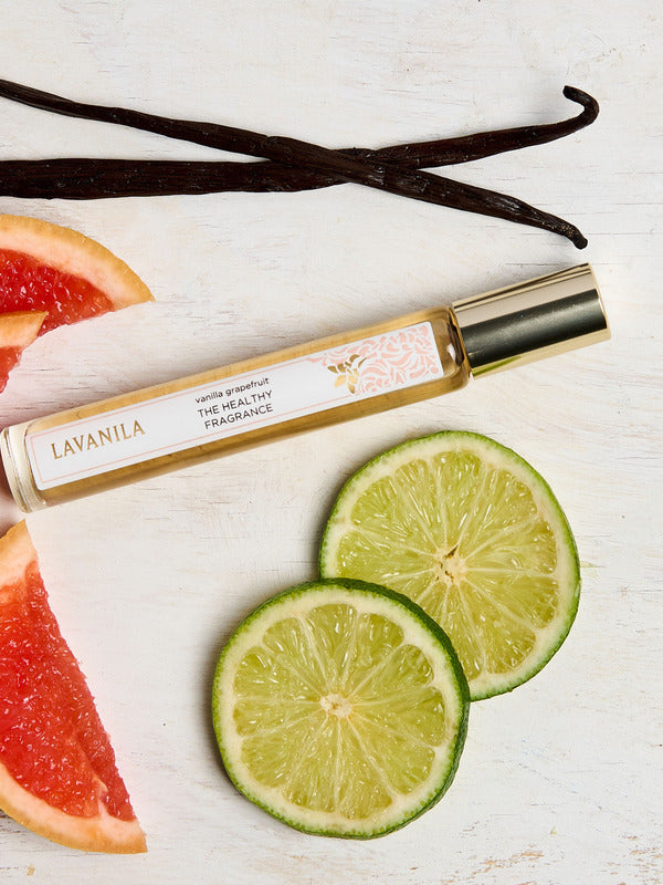 Slim Lavanila Vanilla Grapefruit rollerball perfume bottle with a gold cap lying diagonally on pale wood beside lime slices, pink grapefruit wedges and dark vanilla pods.