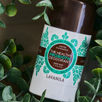 Lavanila Eucalyptus Vanilla probiotic deodorant with a dark brown cap and turquoise-green botanical label lying diagonally on dark wood, framed by green leafy branches.