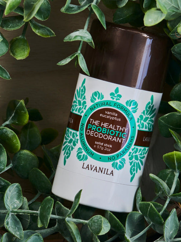 Lavanila Eucalyptus Vanilla probiotic deodorant with a dark brown cap and turquoise-green botanical label lying diagonally on dark wood, framed by green leafy branches.