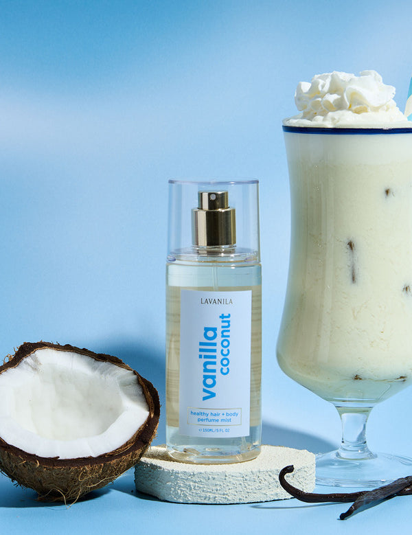 Lavanila Vanilla Coconut Hair and Body Mist arranged with fresh coconut, vanilla pods and a creamy coconut drink on blue.