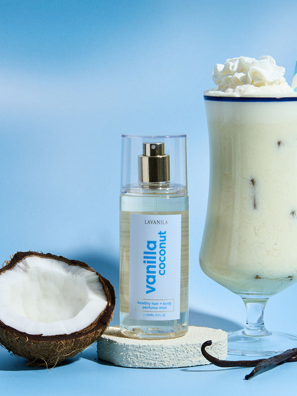 Lavanila Vanilla Coconut Hair and Body Mist arranged with fresh coconut, vanilla pods and a creamy coconut drink on blue.