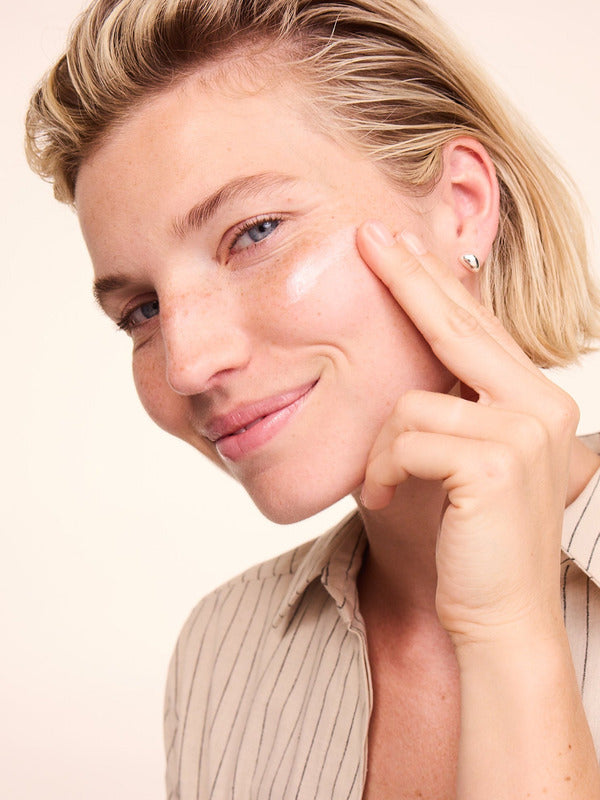 Woman gently applying Lavera Basis Sensitiv Calming Night Cream to her cheek against a soft peach background.