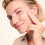 Woman smiling while applying a white streak of Lavera Basis Sensitiv Q10 Moisturising Day Cream to her cheek with one fingertip.