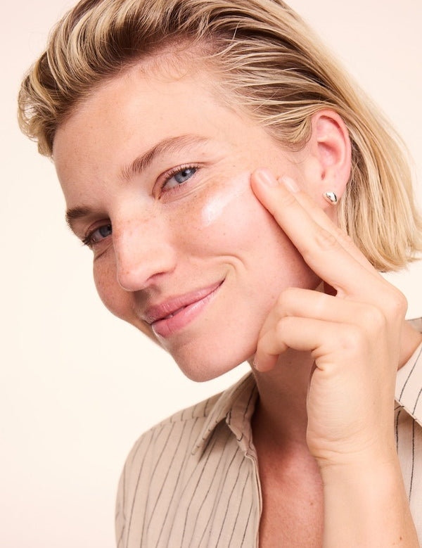 Woman smiling while applying a white streak of Lavera Basis Sensitiv Q10 Moisturising Day Cream to her cheek with one fingertip.