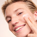 Smiling woman applying Lavera Basis Sensitiv Q10 Night Cream to her cheek against a soft peach background.