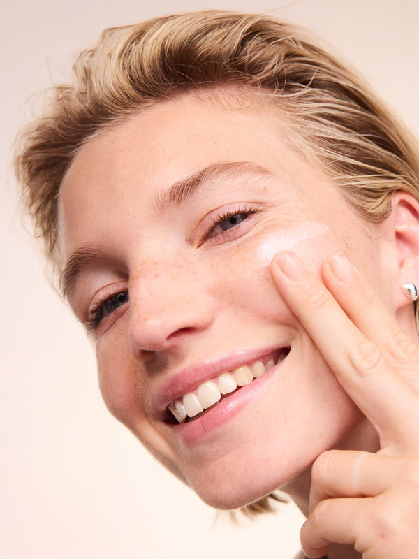 Smiling woman applying Lavera Basis Sensitiv Q10 Night Cream to her cheek against a soft peach background.