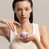 Woman holding a jar of Lavera Firming Day Cream while touching the cream with one fingertip.