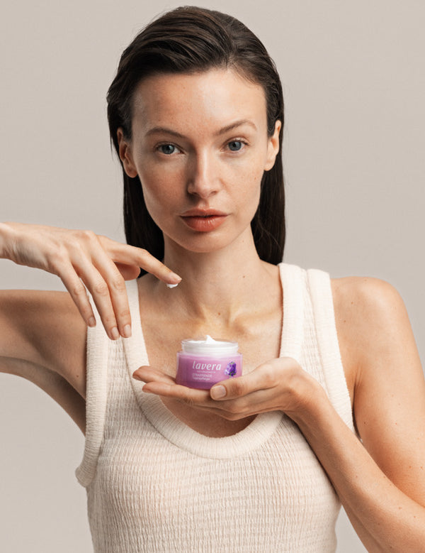 Woman holding a jar of Lavera Firming Day Cream while touching the cream with one fingertip.