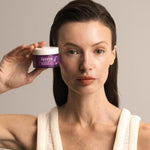 Woman holding a purple jar of Lavera Firming Night Cream beside her face against a warm neutral background.