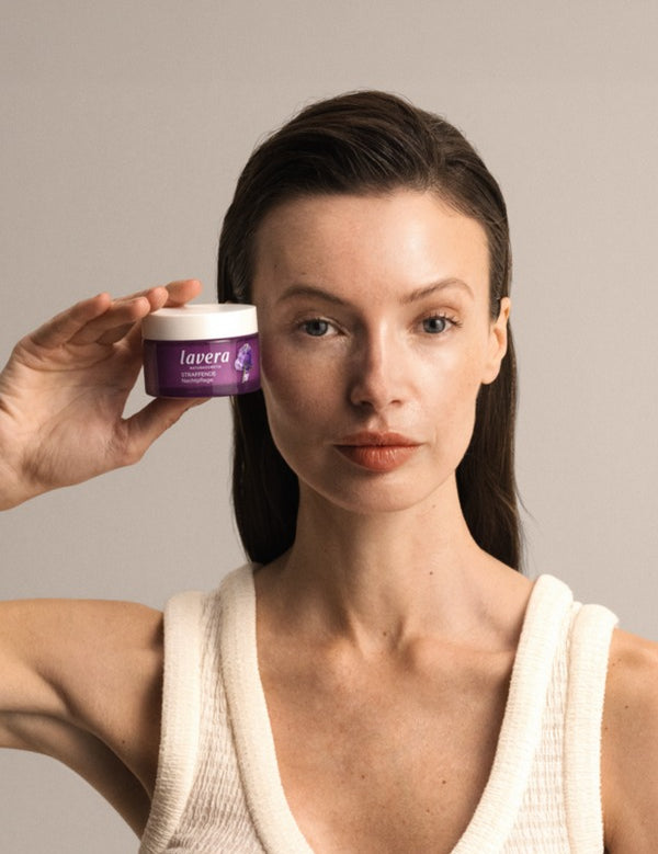 Woman holding a purple jar of Lavera Firming Night Cream beside her face against a warm neutral background.