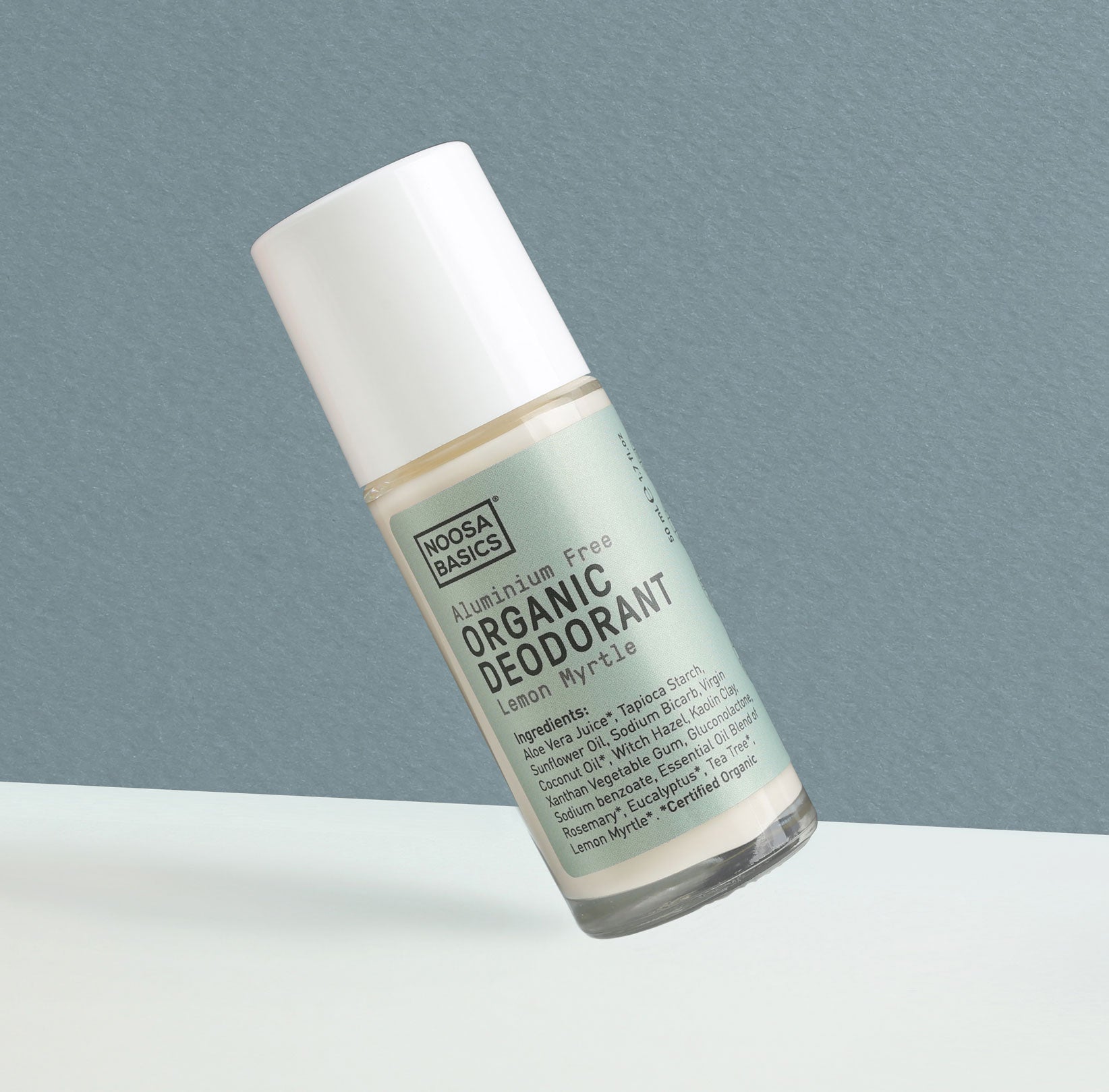 Clear Noosa Basics Lemon Myrtle roll-on deodorant bottle with a pale green label and white cap tilted diagonally against a grey-green background.