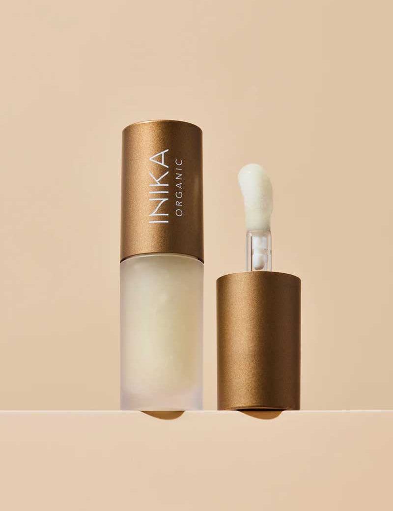 A frosted bottle of INIKA Organic Lip Oil - Collagen + Squalane Clear 5ml with a bronze cap is displayed; the cap stands beside the bottle, exposing a sponge-tip applicator. The backdrop features a soft beige tone.