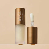 A matte gold tube of INIKA Organic Lip Oil - Collagen + Squalane Clear stands upright with its matching wand applicator, featuring a soft white tip, beside it on a neutral beige background.