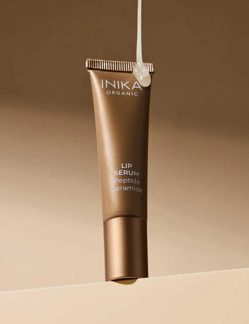 An upright bronze tube of INIKA Organic Tinted Lip Serum - Peptide + Ceramide Clear 10ml stands against a beige background, with a drop of serum above the cap, highlighting its organic nourishing power.