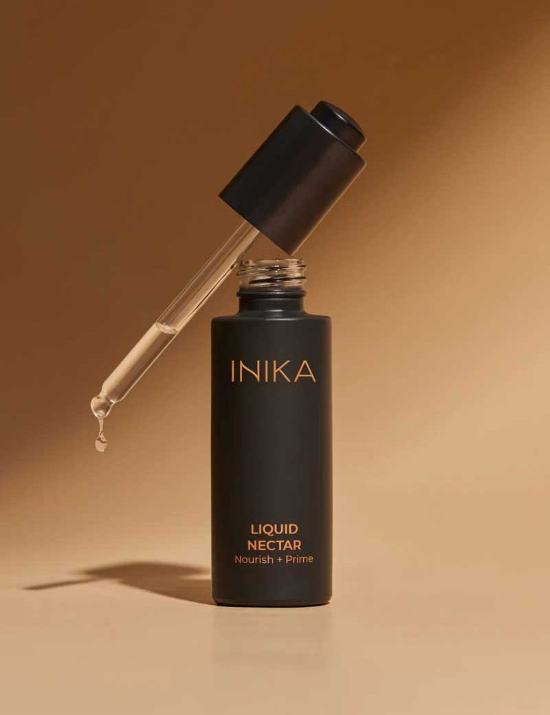 A matte black bottle of INIKA Organic Liquid Nectar Primer 30ml stands upright with an open dropper cap and a visible drop of primer. Set against a warm beige gradient, it highlights this certified organic essential for nourished, primed skin.