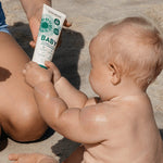 Baby holding a tube of Little Urchin Baby Natural Clear Zinc Sunscreen SPF50+ while seated with a parent on the beach.
