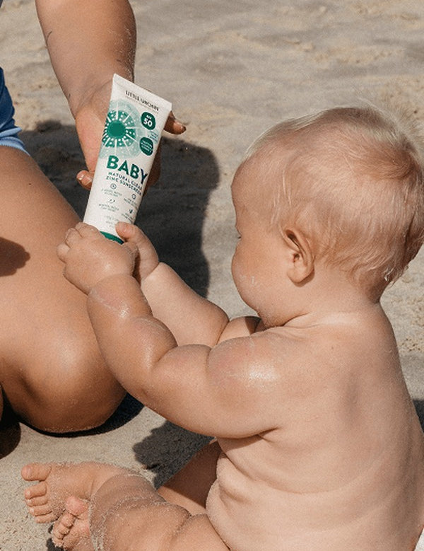 Baby holding a tube of Little Urchin Baby Natural Clear Zinc Sunscreen SPF50+ while seated with a parent on the beach.