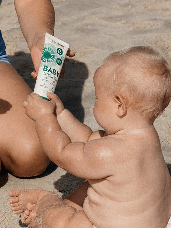 Baby holding a tube of Little Urchin Baby Natural Clear Zinc Sunscreen SPF50+ while seated with a parent on the beach.